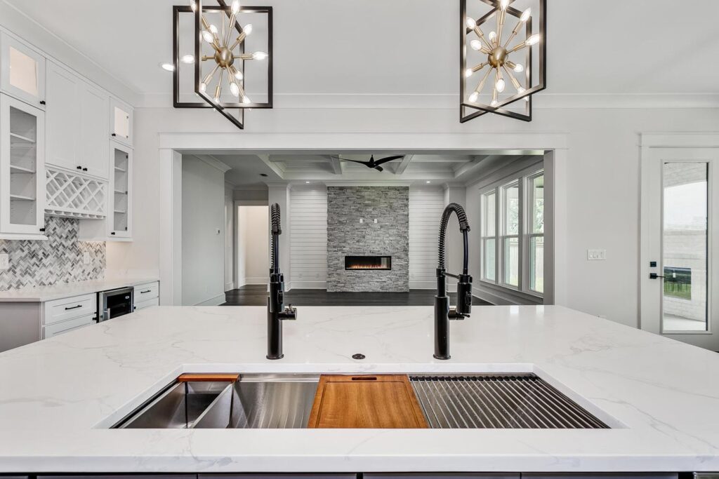 A kitchen island with a dual sink and two chandeliers, looking out onto the living room fireplace